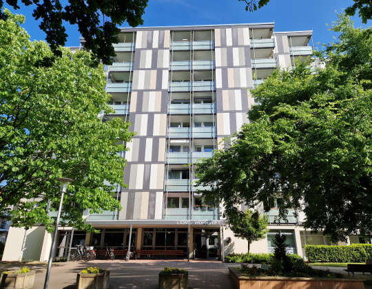 Frontansicht des Wohnstifts. Über dem Eingang ist der Lions Wohnstift Schriftzug zu sehen. Das Gebäude wird von Bäumen umrandet.